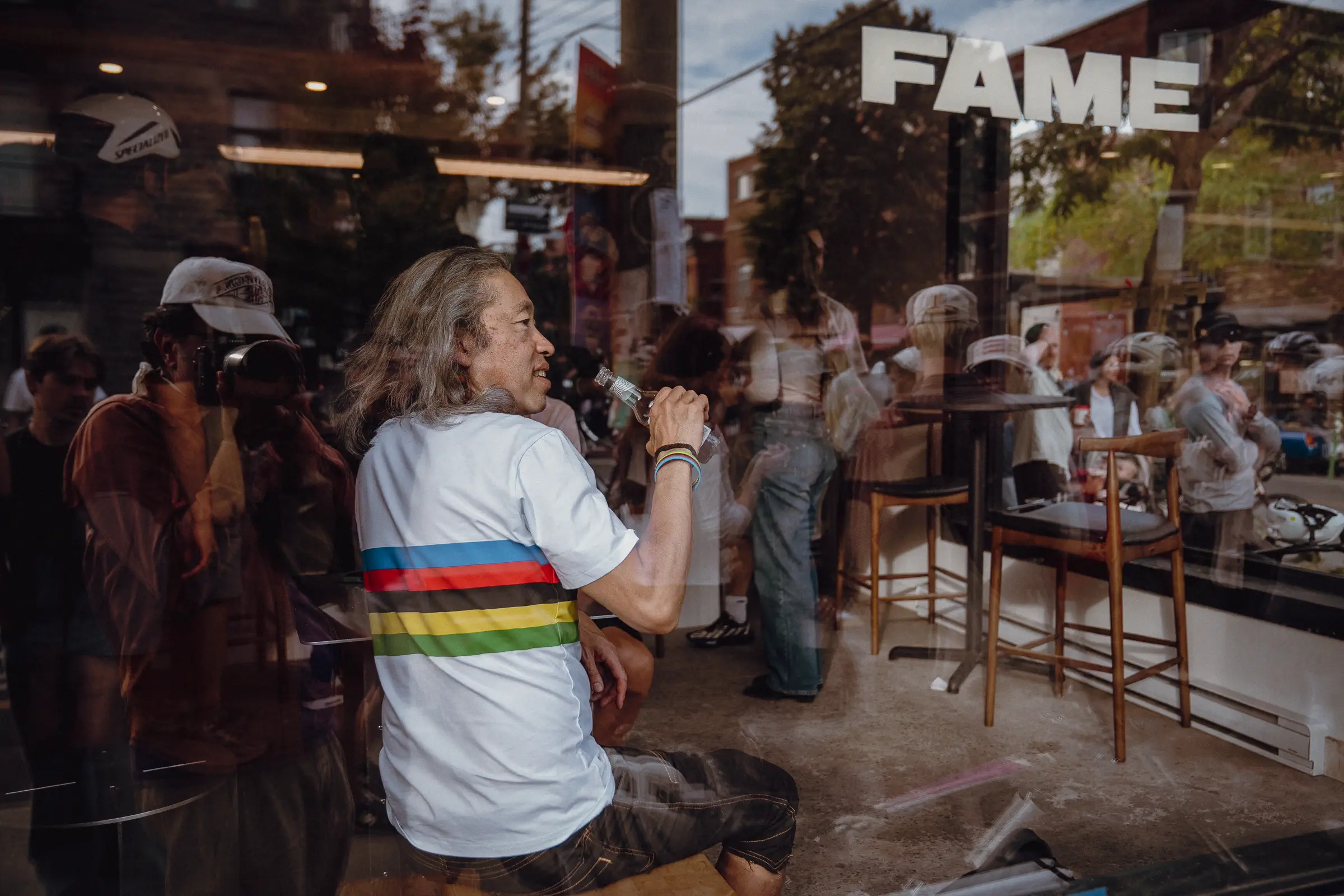 A rainbow-jersey figure appears through reflections in a Montréal café window after the race