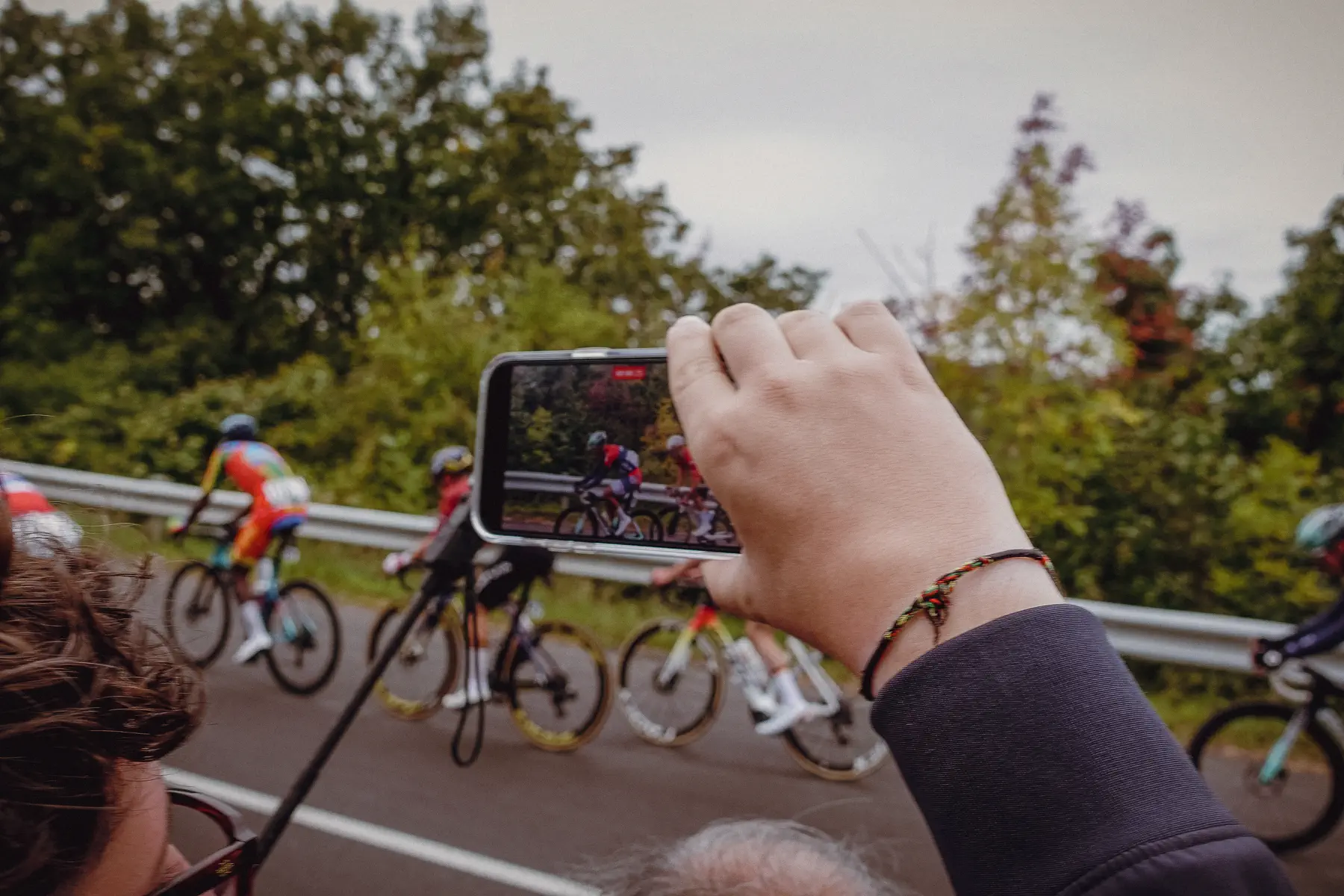 A spectator holds up a phone as the race sweeps past just beyond the barrier
