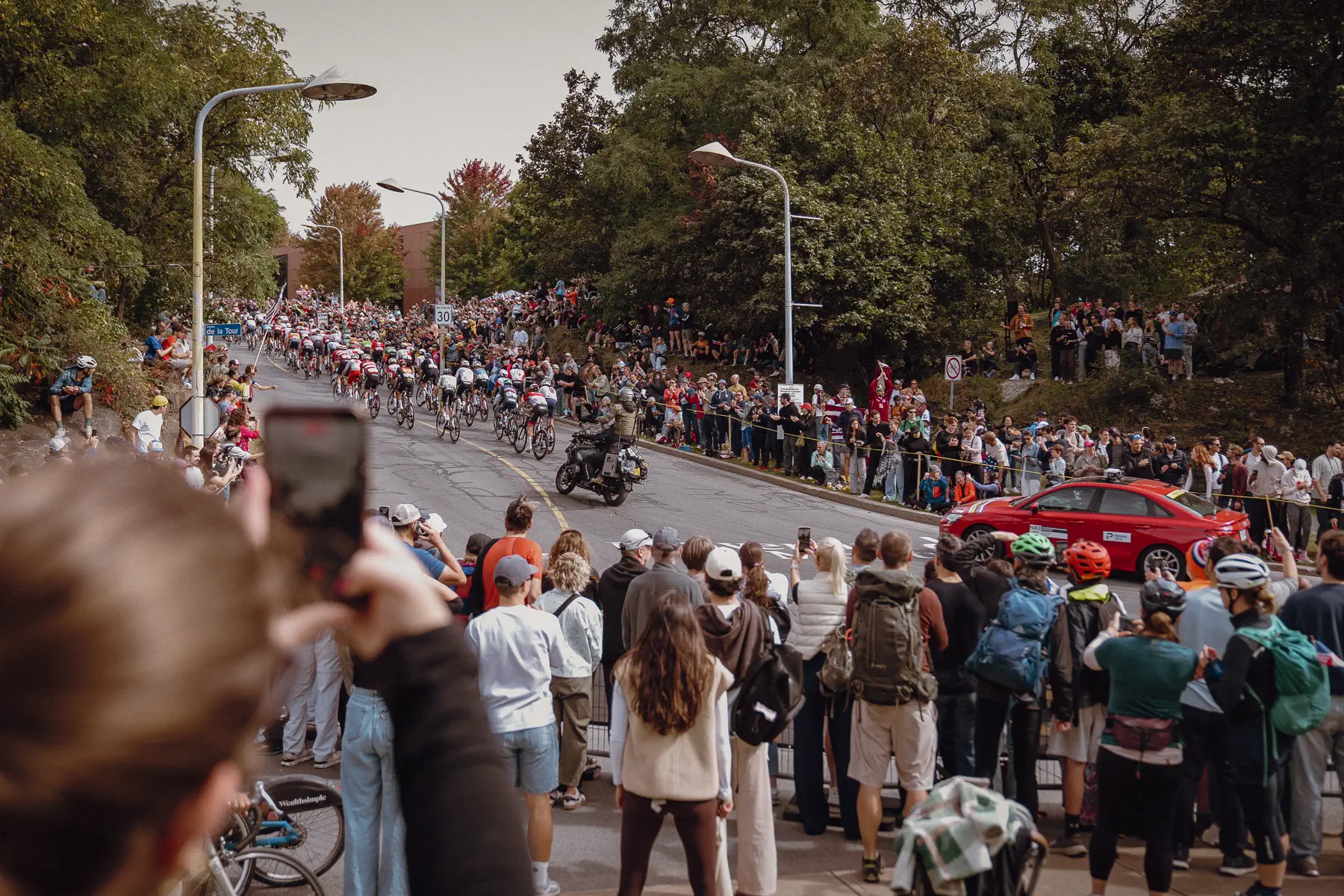 A long line of riders bends through Mount Royal with spectators stacked deep along the course